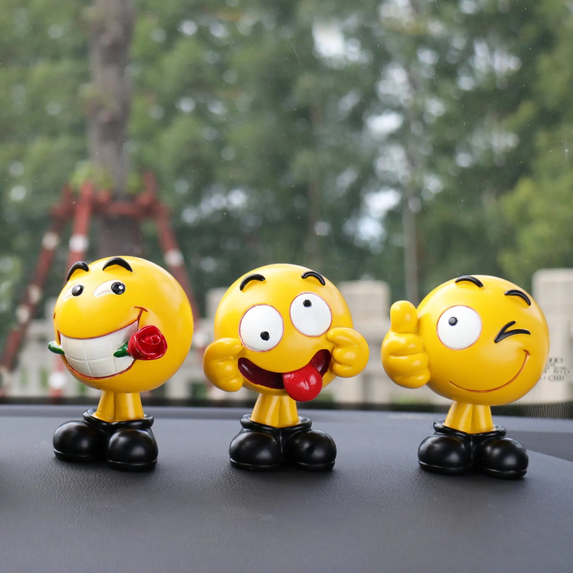 Three yellow smiley face figurines on a car dashboard with a blurred outdoor background.